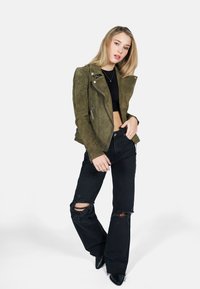 Olijfgroene suède motorjack met ritsen, gecombineerd met zwarte gescheurde flared jeans en puntige zwarte schoenen, tegen een witte achtergrond.