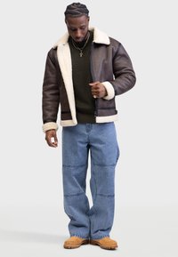 Chaqueta de cuero marrón con cuello y puños de borrego blanco, combinada con un suéter de punto verde y jeans de pierna ancha azul claro. Botas marrones.
