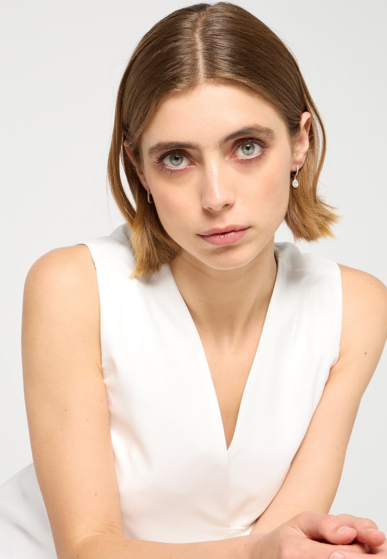 Blouse blanche sans manches avec un décolleté en V, texture lisse et drapé doux. Le modèle a les cheveux courts et raides ainsi que des yeux verts prononcés.