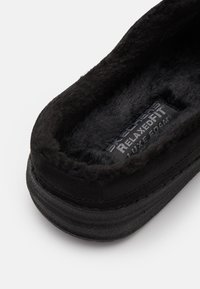 Zwarte pantoffel met een pluche, textuurinterieur en een stevige buitenzool. Het label leest "Skechers Relaxed Fit Luxe Foam."