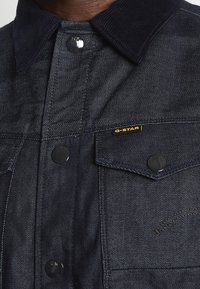 Mörk denimbombare med en kordflöjelskrage, utrustad med metallknappar och en G-STAR-märkning. Texturerat tyg med en strukturerad design.