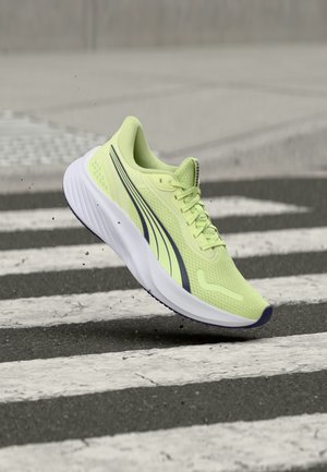 Chaussure de course vert clair avec des bandes noires survolant un passage piéton zébré sur l'asphalte, de petites particules de poussière visibles dans l'air.