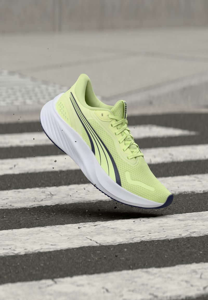 Világoszöld futócipő fekete csíkokkal, amely egy zebrán lebeg aszfalton, apró szennyeződésrészecskék láthatók a levegőben.