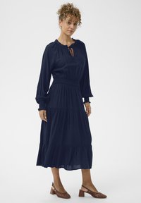 Navyblauw, langärmige jurk met een smocktaille, rimpeldetails en een gelaagde rok. Draag met bruine, open teen, gekruiste hakken.