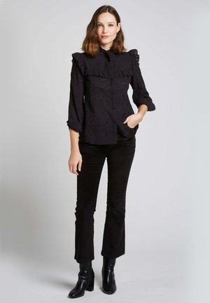 Chemise noire à boutons avec texture florale et col à volants, associée à un pantalon noir évasé et des bottines à talons.