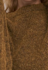 Zbliżenie na osobę w brązowym swetrze o puszystej fakturze, ukazujące szczegóły tkaniny oraz część szyi i włosów.