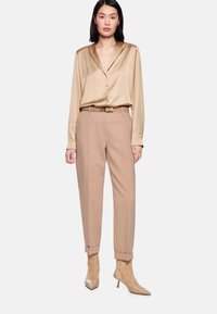 Satinbeige Bluse mit langen Ärmeln und Reverskragen, kombiniert mit hoch taillierten taupefarbenen Hosen. Spitz zulaufende beige Absatzschuhe vervollständigen den Look.