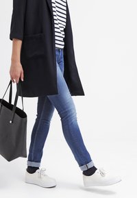 Svart lång kappa över en randig skjorta, kombinerad med blå jeans och vita sneakers. En svart tote-väska h ålls i ena handen.