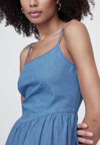 Traper haljina s tankim spaghetti naramenicama, pripijenim gornjim dijelom i suknjom koja se širi. Svijetloplave boje i glatke teksture s naborima u struku.