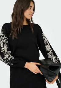 Zwarte pullover trui met lange mouwen, voorzien van witte bloemenborduurwerk op de mouwen. Model houdt een zwarte leren handtas vast.
