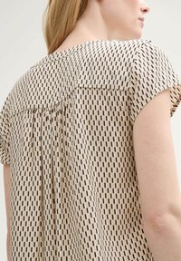 Haut beige clair présentant un motif géométrique noir, avec des manches courtes, un tissu doux et un design à dos plissé.