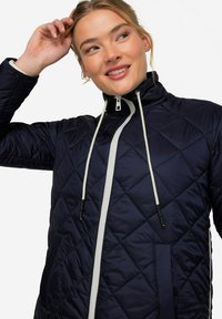 Marinblå quiltad jacka med hög krage, vit dragkedja framtill och dragsko. Har sidfickor och en texturerad, vadderad yta.
