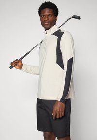 Lys beige langermet skjorte med svarte detaljer, glidelås-kollarfeste og geometrisk design. Kombinert med svarte shorts. Golfkølle holdt i høyre hånd.