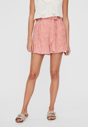 Frau trägt rosa strukturierte High-Waist-Shorts mit Bindegürtel, weiße ärmellose Bluse und beigefarbene Slipper-Sandalen mit Schnallen.
