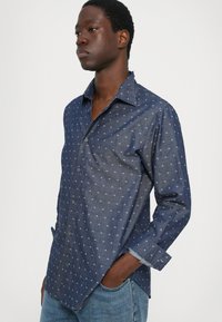 Camisa de mezclilla en azul oscuro con un patrón de estrellas blancas, con cuello clásico, cierre de botones en el frente y mangas largas con puños de botón.