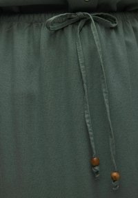 Ceinture élastique en tissu vert avec deux perles en bois. Texture douce et design froncé. Comprend une fermeture à bouton en haut.