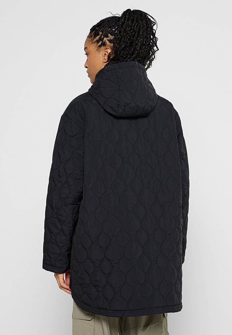 Gestepptes schwarzes Kapuzenjackett mit schmalem Schnitt. Hat eine strukturierte Oberfläche mit einem geometrischen Muster und seitlichen Taschen.