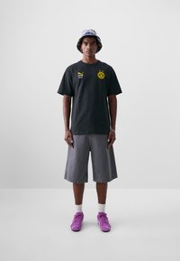 Svart T-skjorte med gul logo, grå denimshorts, lilla joggesko og en rutete bøttehatt. Enkle, uformelle klær med en avslappet passform.