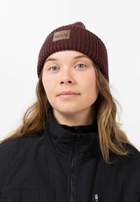 Maroon ribbed knit beanie with a woven label on the front; worn with a black zip-up jacket; smooth texture and fitted shape.