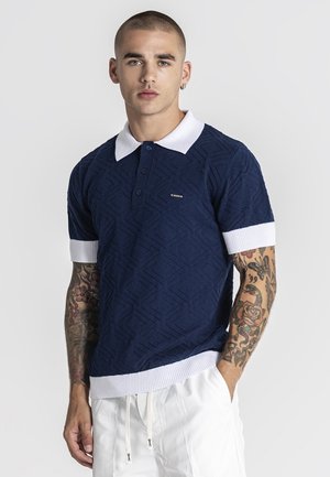 Giovane uomo con capelli corti e tatuaggi su entrambe le braccia, indossa una polo blu navy strutturata con colletto e polsini bianchi, pantaloni bianchi, in piedi davanti a una parete semplice.