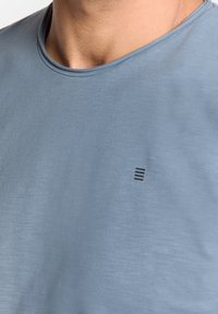 Lichtblauwe T-shirt met korte mouwen van zacht materiaal, voorzien van een ronde hals. Heeft een klein zwart gestreept logo op de borst.
