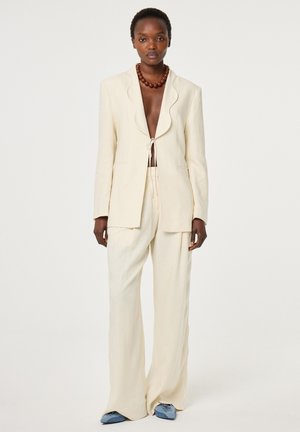 Lange vrouw die een crèmekleurig open colbert draagt met bijpassende brede broekspijpen, blauwe puntschoenen en een dikke bruine kralenketting.