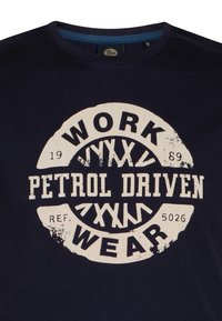 Námořnická modrá bavlněná tričko s velkým kulatým grafickým prvkem obsahujícím text "WORK PETROL DRIVEN WEAR" v béžovém tisku s opotřebenými akcenty.