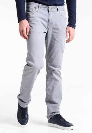 Uomo che indossa jeans grigio chiaro e sneakers blu navy, in piedi con le mani rilassate lungo i fianchi davanti a uno sfondo bianco.