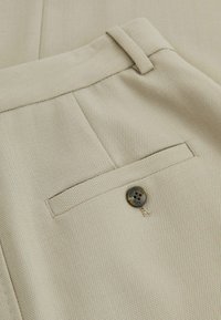 Beige neulehousut, joissa on takatasku ruskealla napilla kiinnitettävänä. Kangas on tekstuuriltaan mielenkiintoinen ja siinä on hienovarainen kuvio.