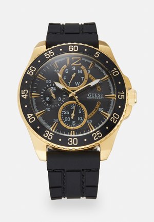 Gold-tone Guess wristwatch with black dial, three subdials, gold markers, and black silicone strap showing 10:09:33 time.