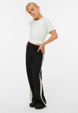 Witte t-shirt met korte mouwen gecombineerd met zwarte broek met crèmekleurige zijstrepen; model poseert zelfverzekerd met de handen in de zakken.