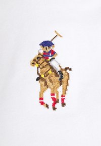 Polo speler met een blauw helm en rood shirt, rijdend op een beige paard met rode beenverbanden, houdt een opgeheven polo mallet vast tegen een witte stoffen achtergrond.