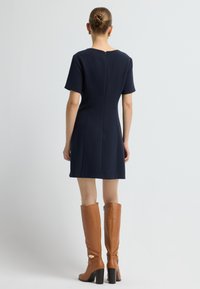 Marineblauwe jurk met korte mouwen, een ronde halslijn en een rits aan de achterkant, gecombineerd met kniehoge bruine leren laarzen met chunky hakken en gespsluiting.
