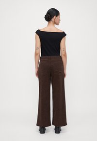 Vrouw met een bun, gekleed in een zwarte off-shoulder top, bruine broek met luipaardprint en zwarte hakken, staand met de rug naar de kijker.