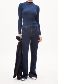 Haut bleu marine à manches longues côtelé associé à un jean évasé bleu foncé. Chaussures argentées réfléchissantes et une veste noire tenue dans une main.