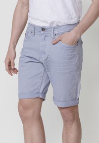 Vīrietis, kurš valkā gaiši zilos īsos šortus ar salocītām malām un baltu T-kreklu, ar vienu roku kabatā uz vienkrāsaina fona.