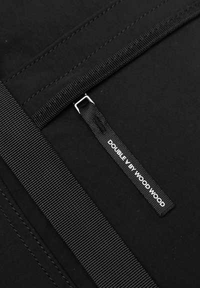 Sac en tissu noir avec une texture lisse, des coutures horizontales, une poche zippée et une étiquette affichant "DOUBLE BY WOOD WOOD."