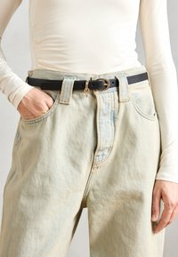 Cinturón de cuero negro con una hebilla circular dorada y un charm, llevado sobre unos pantalones vaqueros de tiro alto de color azul claro con una textura deslavada.