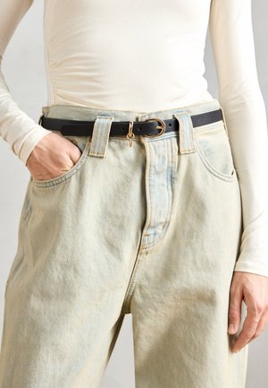 Čierny kožený opasok so zlatou kruhovou prackou a príveskom, nosený cez svetlomodré vysoké rifľové nohavice s vyblednutou textúrou.