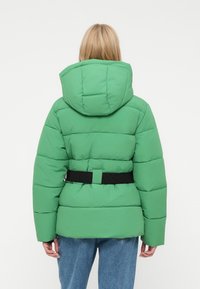 Zelená puffa bunda s kapucňou, s opaskom v páse a prešívanými časťami. Materiál sa zdá byť hladký a mierne lesklý.