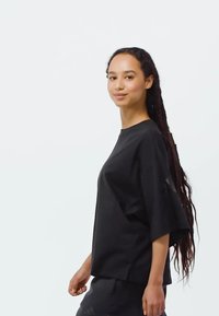 Løs pasform, sort t-shirt, korte ærmer, rund halsudskæring, blød tekstur, sideslids, bæres med matchende mørke bukser, model står foran en hvid baggrund.