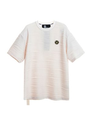 Joven de pie con una camisa beige de manga corta con textura, pantalones cortos azul marino con cordones, calcetines blancos y zapatos beige toscos sin cordones.