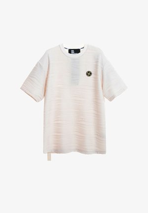 Joven de pie con una camisa beige de manga corta con textura, pantalones cortos azul marino con cordones, calcetines blancos y zapatos beige toscos sin cordones.