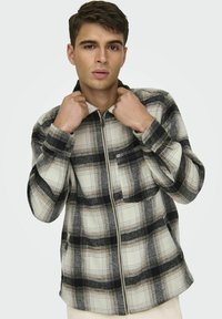 Koszula w kratę z zamkiem w kolorach czarnym, białym i beżowym; ma kołnierzyk i mankiety zapinane na guziki, wykonana z miękkiego materiału o wzorze w kratę.
