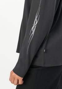 Nærbilde av en persons arm iført en mørk langermet skjorte med et sølvfarget tribal flammedesign på ermene nær håndleddet.