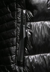 Chaleco acolchado negro con textura brillante y un cierre lateral. Material reflectante contrasta con elementos mate.