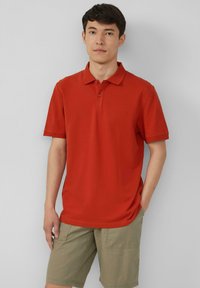 Rode polo met een klassieke kraag, korte mouwen en een drieknopssluiting, gecombineerd met khaki shorts. Zachte textuur, minimalistisch ontwerp.