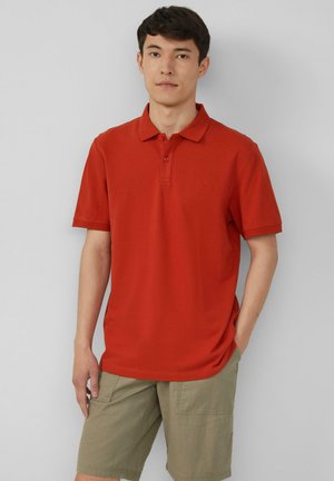 Rotes Polo-Shirt mit klassischem Kragen, kurzen Ärmeln und einer Drei-Knopf-Leiste, kombiniert mit khakifarbenen Shorts. Weicher Stoff, minimalistisches Design.