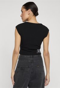 Tumeses juuksepunus naine, kes kannab musta varrukateta lühikest toppi ja kõrge vöökohaga tuhmiks kulunud musta teksapükse, vaadates seljaga otse lihtsa tausta poole.