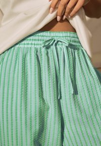 Vrouw tilt een wit shirt op, waarbij ze haar middel en de tailleband van een groen en lichtblauw gestreepte broek met een gestrikte koord laat zien.